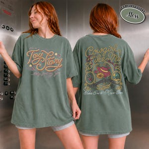 K&ouml;nnte beinhalten: Ein moosgr&uuml;nes Kurzarm-T-Shirt mit einer Vordergrafik, die den Text Toy Story in einer seilartigen Schrift zeigt. Die R&uuml;ckseite zeigt den Text Cowgirl Like Me und Takes One to Know One mit Illustrationen von einem Hut, Seil und Blumen.