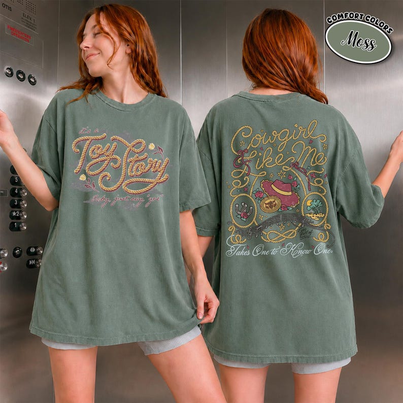 K&ouml;nnte beinhalten: Ein moosgr&uuml;nes Kurzarm-T-Shirt mit einer Vordergrafik, die den Text Toy Story in einer seilartigen Schrift zeigt. Die R&uuml;ckseite zeigt den Text Cowgirl Like Me und Takes One to Know One mit Illustrationen von einem Hut, Seil und Blumen.