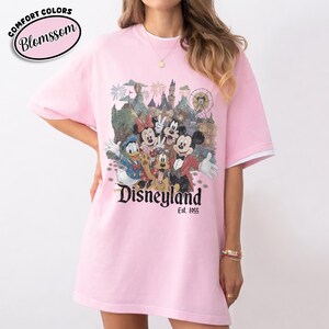 Może przedstawiać: R&oacute;żowa koszulka z grafiką Disneyland z Myszką Miki, Minnie, Kaczorem Donaldem, Goofym i Pluto. Koszulka ma białe wykończenia na rękawach i napis "Disneyland Est. 1955". W lewym g&oacute;rnym rogu znajduje się napis "Comfort Colors Blomssom".