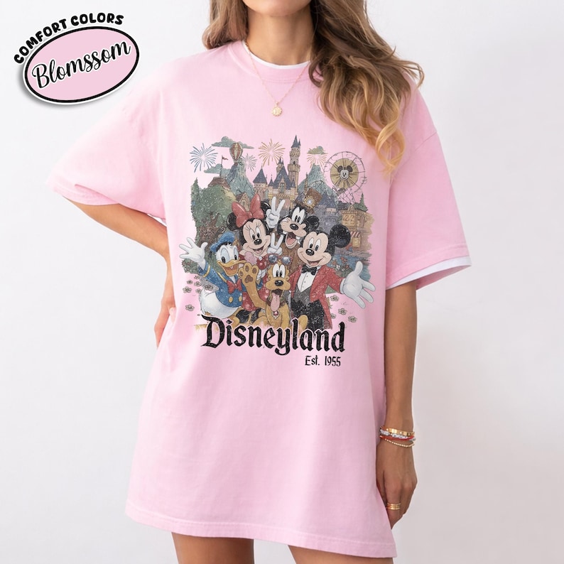 Może przedstawiać: R&oacute;żowa koszulka z grafiką Disneyland z Myszką Miki, Minnie, Kaczorem Donaldem, Goofym i Pluto. Koszulka ma białe wykończenia na rękawach i napis "Disneyland Est. 1955". W lewym g&oacute;rnym rogu znajduje się napis "Comfort Colors Blomssom".