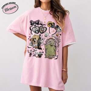 May include: A light pink short-sleeved t-shirt featuring a graphic print of characters from The Nightmare Before Christmas, including Jack Skellington, Sally, and Oogie Boogie, along with a spiderweb, bats, and a headband with ears.
