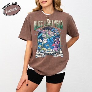 May include: A brown t-shirt featuring a graphic of Buzz Lightyear from Toy Story. The design includes the text "Buzz Lightyear" above an image of Buzz in flight, with other characters and the text "Disney Laser Blast" below.