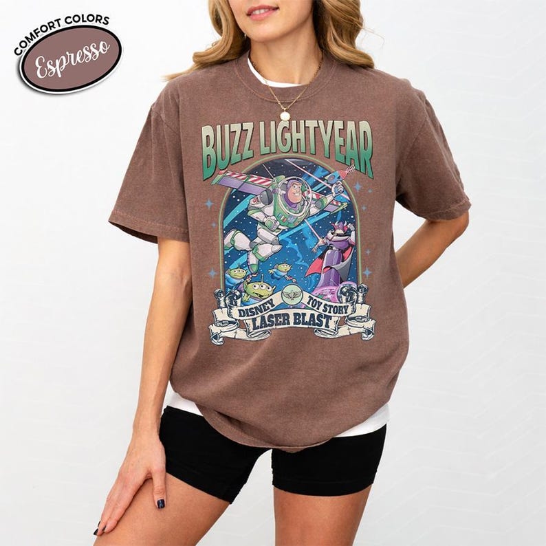 May include: A brown t-shirt featuring a graphic of Buzz Lightyear from Toy Story. The design includes the text "Buzz Lightyear" above an image of Buzz in flight, with other characters and the text "Disney Laser Blast" below.