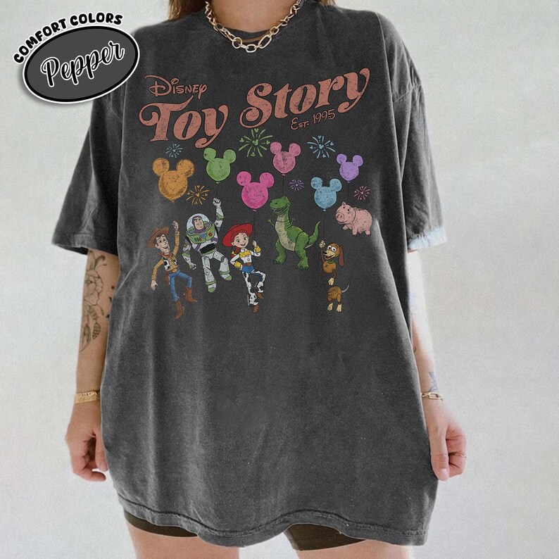 K&ouml;nnte beinhalten: Dunkelgraues T-Shirt mit dem Disney Toy Story Logo und Charakteren. Das Design umfasst Woody, Buzz Lightyear, Jessie, Rex, Hamm und Slinky Dog, mit bunten Mickey Mouse Luftballons. Das T-Shirt ist von der Marke Comfort Colors.