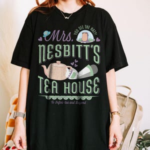 K&ouml;nnte beinhalten: Schwarzes T-Shirt mit einem gr&uuml;nen und wei&szlig;en Grafikdesign mit der Aufschrift "Mrs. Nesbitt's Tea House". Das Design umfasst eine Teekanne, eine Tasse und den Satz "To Infini-tea and Beyond".