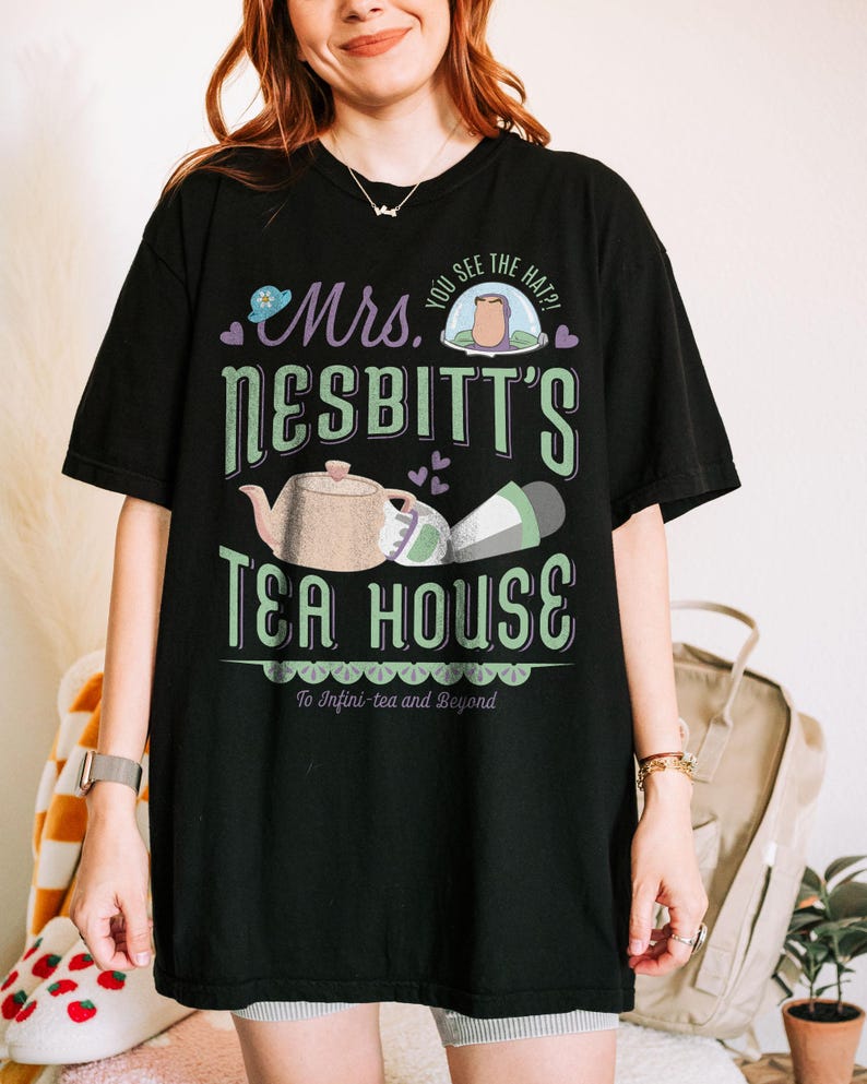 K&ouml;nnte beinhalten: Schwarzes T-Shirt mit einem gr&uuml;nen und wei&szlig;en Grafikdesign mit der Aufschrift "Mrs. Nesbitt's Tea House". Das Design umfasst eine Teekanne, eine Tasse und den Satz "To Infini-tea and Beyond".