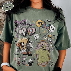 May include: A moss green t-shirt featuring a graphic print of characters and icons from The Nightmare Before Christmas. The design includes Jack Skellington, Sally, Zero, Oogie Boogie, and various holiday-themed elements like bats and candy corn.