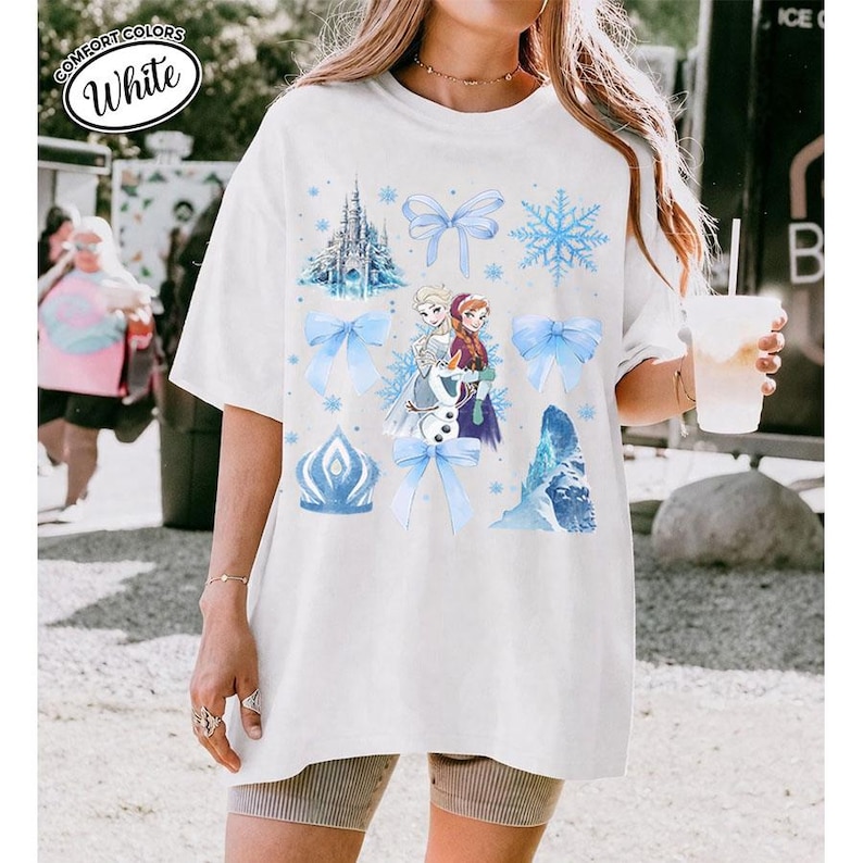Puede incluir: Camiseta blanca Comfort Colors con un dise&ntilde;o de la pel&iacute;cula Frozen. El dise&ntilde;o incluye a Elsa, Anna, copos de nieve, lazos y un castillo en tonos azules.