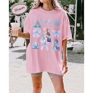 Puede incluir: Una camiseta rosa claro Comfort Colors con un gr&aacute;fico que presenta personajes y elementos de una popular pel&iacute;cula animada. El dise&ntilde;o incluye un castillo, copos de nieve, lazos y la palabra "Blossom".