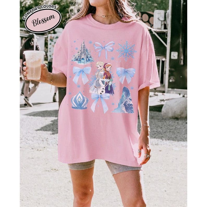 Puede incluir: Una camiseta rosa claro Comfort Colors con un gr&aacute;fico que presenta personajes y elementos de una popular pel&iacute;cula animada. El dise&ntilde;o incluye un castillo, copos de nieve, lazos y la palabra "Blossom".