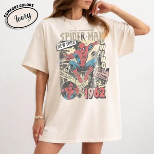 Puede incluir: Camiseta color marfil con un gr&aacute;fico vintage de Spider-Man. El dise&ntilde;o incluye las palabras "The Amazing Spider-Man", "New York" y "1962". El gr&aacute;fico representa a Spider-Man en una pose din&aacute;mica, con un estilo retro de c&oacute;mic.