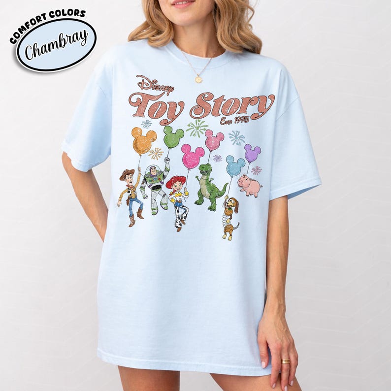 K&ouml;nnte beinhalten: Hellblaues T-Shirt mit dem Disney Toy Story-Logo und Figuren, die ballonf&ouml;rmige Ballons halten. Das Shirt hat einen Rundhalsausschnitt und kurze &Auml;rmel. Der Text "Comfort Colors Chambray" befindet sich oben links.