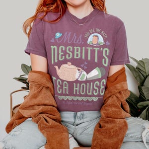 K&ouml;nnte beinhalten: Ein lila T-Shirt mit dem Text "Mrs. Nesbitt's Tea House" in Gr&uuml;n und Wei&szlig;. Das Design beinhaltet eine Teekanne, eine Tasse und den Satz "To Infini-tea and Beyond". Das Shirt wird mit einer braunen Jacke und blauen Jeans getragen.