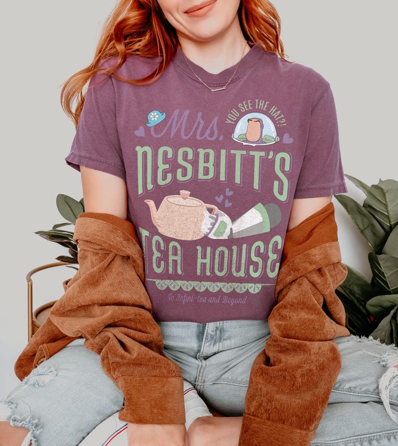 K&ouml;nnte beinhalten: Ein lila T-Shirt mit dem Text "Mrs. Nesbitt's Tea House" in Gr&uuml;n und Wei&szlig;. Das Design beinhaltet eine Teekanne, eine Tasse und den Satz "To Infini-tea and Beyond". Das Shirt wird mit einer braunen Jacke und blauen Jeans getragen.
