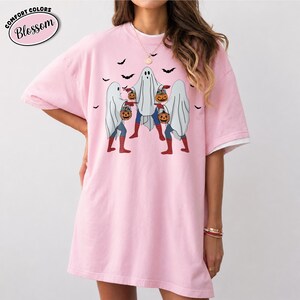 May include: Pale pink Comfort Colors t-shirt featuring a Halloween design. The graphic shows three ghosts carrying pumpkins, with bats overhead. The shirt has the word "Blossom" in a pink oval.