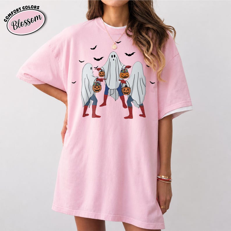 May include: Pale pink Comfort Colors t-shirt featuring a Halloween design. The graphic shows three ghosts carrying pumpkins, with bats overhead. The shirt has the word "Blossom" in a pink oval.