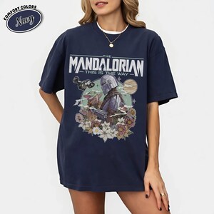 May include: Navy blue t-shirt featuring a graphic of The Mandalorian with Grogu, surrounded by flowers, and the text "THE MANDALORIAN THIS IS THE WAY". The shirt has a relaxed fit and the words "COMFORT COLORS" and "Navy" are in a circle.