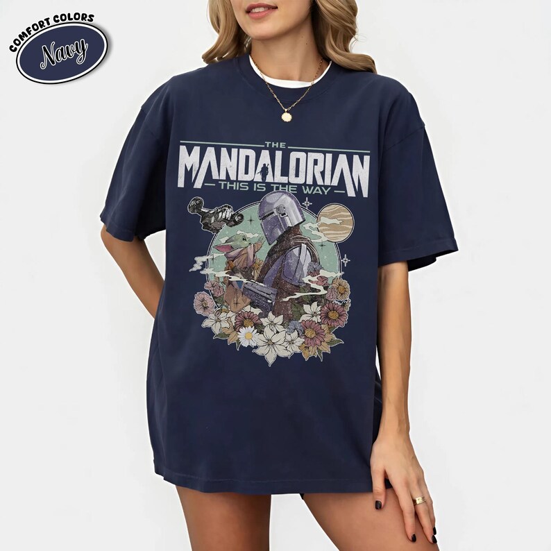 May include: Navy blue t-shirt featuring a graphic of The Mandalorian with Grogu, surrounded by flowers, and the text "THE MANDALORIAN THIS IS THE WAY". The shirt has a relaxed fit and the words "COMFORT COLORS" and "Navy" are in a circle.