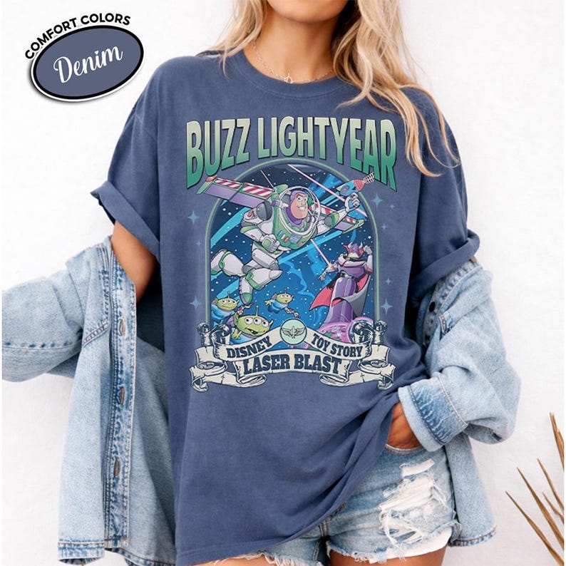 May include: Denim blue t-shirt featuring a graphic of Buzz Lightyear from Disney's Toy Story. The design includes the text "BUZZ LIGHTYEAR", "DISNEY TOY STORY LASER BLAST", and images of the characters. The shirt has a crew neck and short sleeves.
