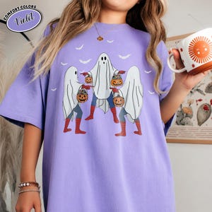 May include: A lilac t-shirt with a Halloween design of three ghost figures dressed as superheroes, each carrying a pumpkin. The shirt has the text "Comfort Colors Violet" in a circular design.