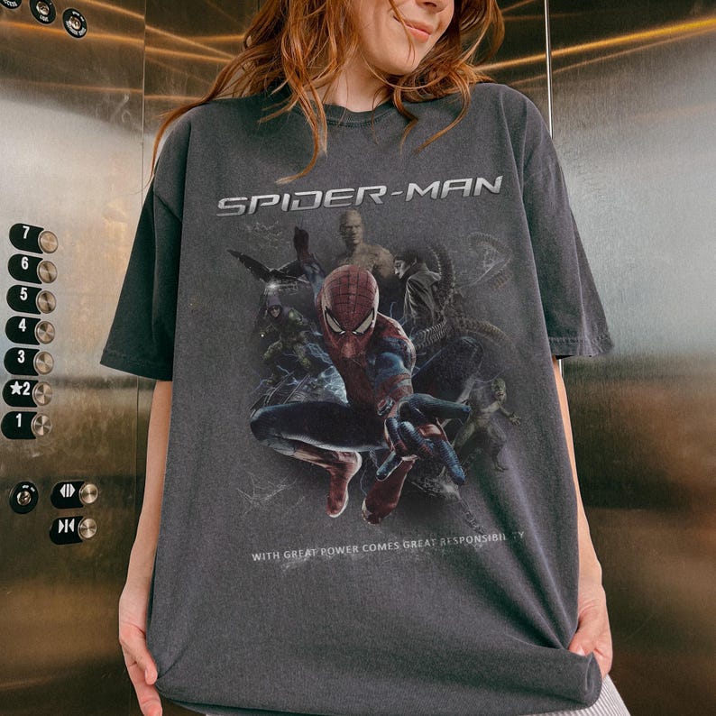 May include: Dark gray Spider-Man t-shirt featuring a graphic of the superhero in a dynamic pose, surrounded by other characters. The text "SPIDER-MAN" is displayed above the image, with the phrase "WITH GREAT POWER COMES GREAT RESPONSIBILITY" below.
