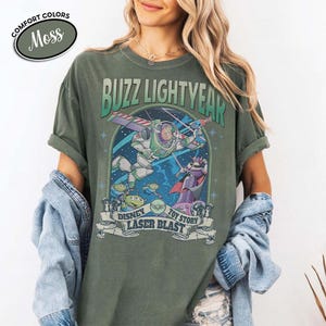May include: Moss green t-shirt featuring a graphic of Buzz Lightyear from Toy Story. The design includes Buzz in flight, aliens, and Zurg, with the text "BUZZ LIGHTYEAR" and "DISNEY TOY STORY LASER BLAST". The shirt has rolled-up sleeves.