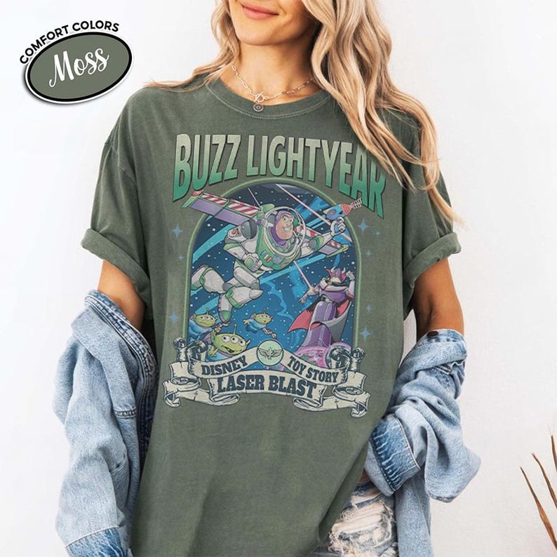 May include: Moss green t-shirt featuring a graphic of Buzz Lightyear from Toy Story. The design includes Buzz in flight, aliens, and Zurg, with the text "BUZZ LIGHTYEAR" and "DISNEY TOY STORY LASER BLAST". The shirt has rolled-up sleeves.