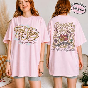 K&ouml;nnte beinhalten: Ein hellrosa T-Shirt mit einem Frontaufdruck, der den Text "it's a Toy Story Baby, just say "yes"" in seilartiger Schrift zeigt. Die R&uuml;ckseite zeigt ein Western-Motiv mit dem Text "Cowgirl like Me" und "Takes One to Know One".