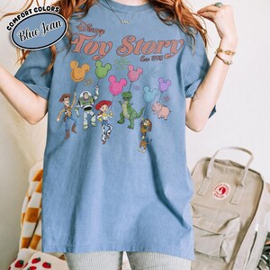 K&ouml;nnte beinhalten: Blaues Jeans-T-Shirt mit dem Disney Toy Story-Logo und Charakteren. Die Grafik zeigt Woody, Buzz Lightyear, Jessie, Rex, Hamm und Slinky Dog, die Mickey Mouse-f&ouml;rmige Ballons halten. Das Shirt hat ein Comfort Colors-Label.