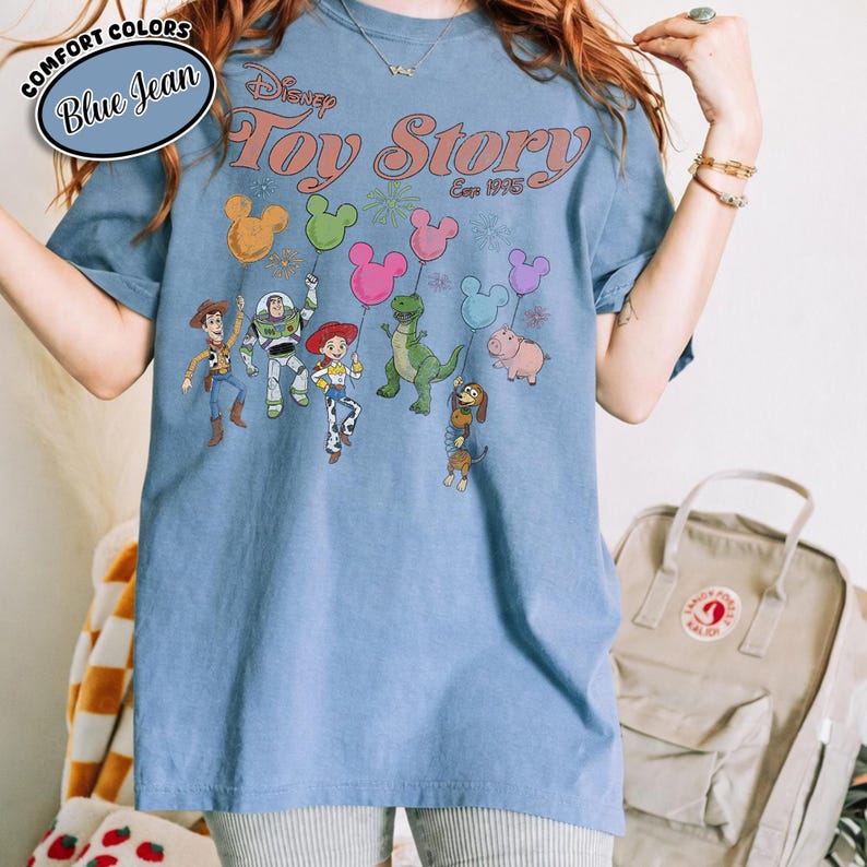 K&ouml;nnte beinhalten: Blaues Jeans-T-Shirt mit dem Disney Toy Story-Logo und Charakteren. Die Grafik zeigt Woody, Buzz Lightyear, Jessie, Rex, Hamm und Slinky Dog, die Mickey Mouse-f&ouml;rmige Ballons halten. Das Shirt hat ein Comfort Colors-Label.