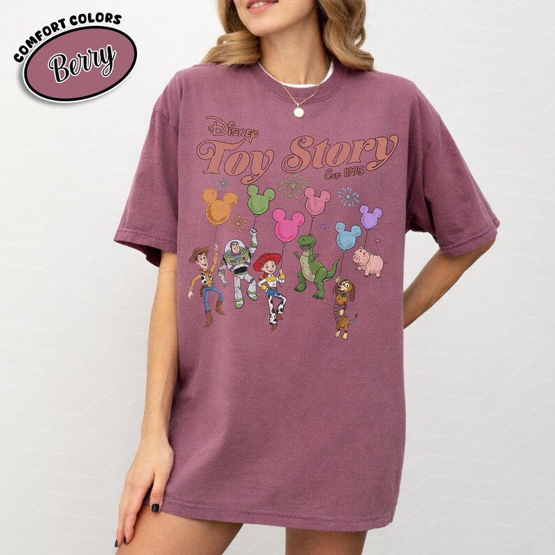 K&ouml;nnte beinhalten: Beerenfarbenes T-Shirt mit dem Disney Toy Story-Logo und -Charakteren. Die Grafik zeigt Woody, Buzz Lightyear, Jessie, Rex, Hamm, Slinky Dog und Ballons. Das Shirt hat die Worte "Comfort Colors" und "Berry" in einem Kreis.