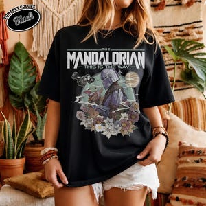 May include: Black t-shirt featuring a graphic of The Mandalorian and Grogu surrounded by flowers. The text reads "THE MANDALORIAN" above "THIS IS THE WAY". The shirt has a crew neck and short sleeves.