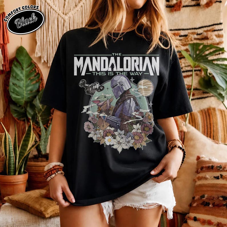 May include: Black t-shirt featuring a graphic of The Mandalorian and Grogu surrounded by flowers. The text reads "THE MANDALORIAN" above "THIS IS THE WAY". The shirt has a crew neck and short sleeves.