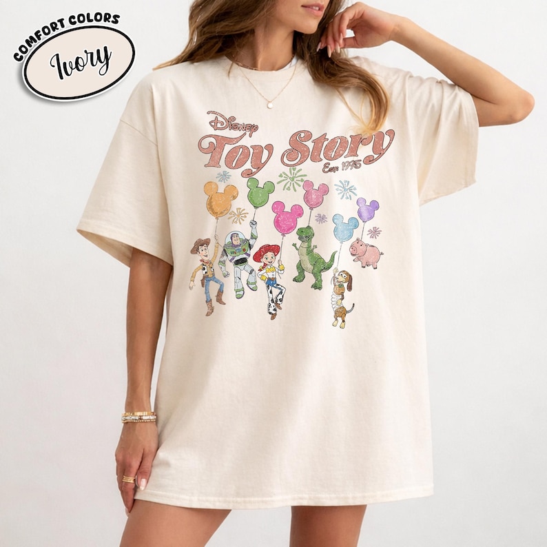 K&ouml;nnte beinhalten: Elfenbeinfarbenes T-Shirt mit einem Aufdruck von Disney's Toy Story-Charakteren, die Ballons halten. Der Text "Disney Toy Story Est. 1995" ist &uuml;ber den Charakteren aufgedruckt. Das Shirt hat eine lockere Passform und die Worte "Comfort Colors" sind oben links aufgedruckt.