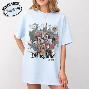 Może przedstawiać: Jasnoniebieska koszulka z grafiką Disneyland z Myszką Miki, Myszka Minnie, Kaczorem Donaldem, Goofym i Pluto. Grafika zawiera zamek, fajerwerki i diabelski młyn. Koszulka ma napis "Disneyland Est. 1955" oraz słowa "Comfort Colors Chambray".
