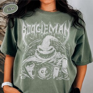 K&ouml;nnte beinhalten: Moosgr&uuml;nes T-Shirt mit einem wei&szlig;en, verblassten Aufdruck des Boogieman-Charakters aus Nightmare Before Christmas. Das Design zeigt das Gesicht der Figur, das Wort "BOOGIEMAN" und andere Gesichter von Charakteren. Das Shirt ist von der Marke Comfort Colors.