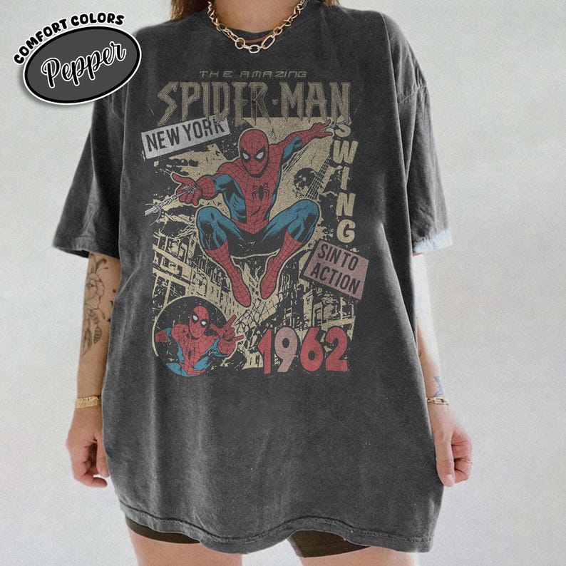 May include: Dark gray t-shirt featuring a vintage Spider-Man graphic. The design includes the text "THE AMAZING SPIDER-MAN", "NEW YORK", and "1962". The shirt has a distressed look and a round neckline.
