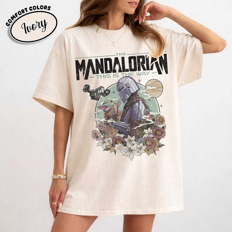 May include: Ivory-colored t-shirt featuring a graphic of The Mandalorian and Grogu surrounded by flowers. The text "THE MANDALORIAN - THIS IS THE WAY" is printed above the illustration. The shirt has short sleeves and a crew neck.