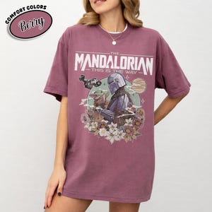 May include: Berry-colored t-shirt featuring a graphic of The Mandalorian with Grogu, surrounded by flowers. The text "THE MANDALORIAN - THIS IS THE WAY" is printed above the image. The shirt has short sleeves and a relaxed fit.