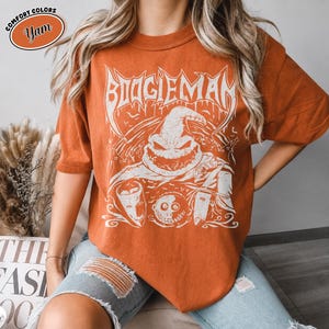 K&ouml;nnte beinhalten: Orangefarbenes T-Shirt mit einem wei&szlig;en Aufdruck der Figur Boogieman. Das Design zeigt das Wort "BOOGIEMAN" in einer stilisierten Schriftart &uuml;ber dem Gesicht der Figur. Das Shirt ist eine Komfortfarbe und hat kurze &Auml;rmel.