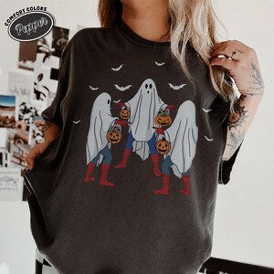 May include: Dark grey t-shirt with a Halloween graphic. The design depicts three ghosts dressed as superheroes, carrying jack-o'-lanterns. Bats fly above the ghosts. The shirt has the text "Comfort Colors Pepper" in a circle at the top left.