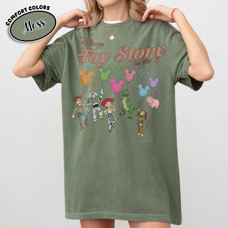 K&ouml;nnte beinhalten: Moosgr&uuml;nes T-Shirt mit dem Disney Toy Story-Logo und Figuren, die ballonf&ouml;rmige Mickey Mouse-K&ouml;pfe halten. Das Shirt hat einen Vintage-Look mit dem Text "Est. 1995". Das Shirt ist eine Komfortfarbe.