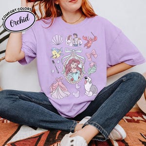 May include: A light purple t-shirt with a graphic featuring Ariel from The Little Mermaid, along with other characters and elements from the movie. The shirt has the words "Comfort Colors" and "Orchid" printed on it.