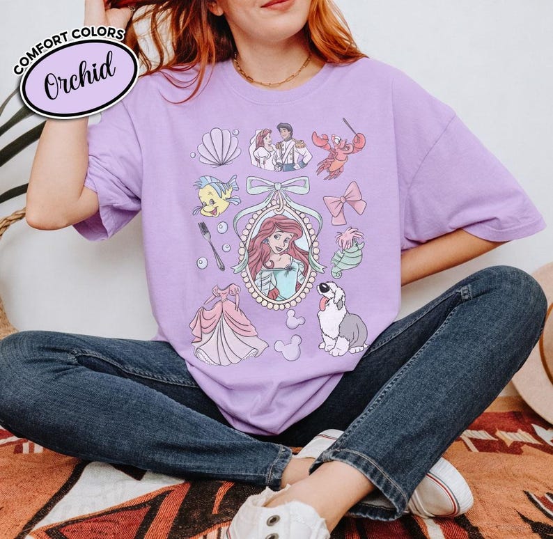 May include: A light purple t-shirt with a graphic featuring Ariel from The Little Mermaid, along with other characters and elements from the movie. The shirt has the words "Comfort Colors" and "Orchid" printed on it.