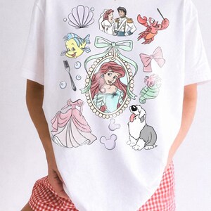 May include: White t-shirt with a colorful graphic featuring characters and elements from a popular animated movie. The design includes a princess, a crab, a fish, and other related imagery. The shirt has the words "Comfort Colors" and "White" in a circle.