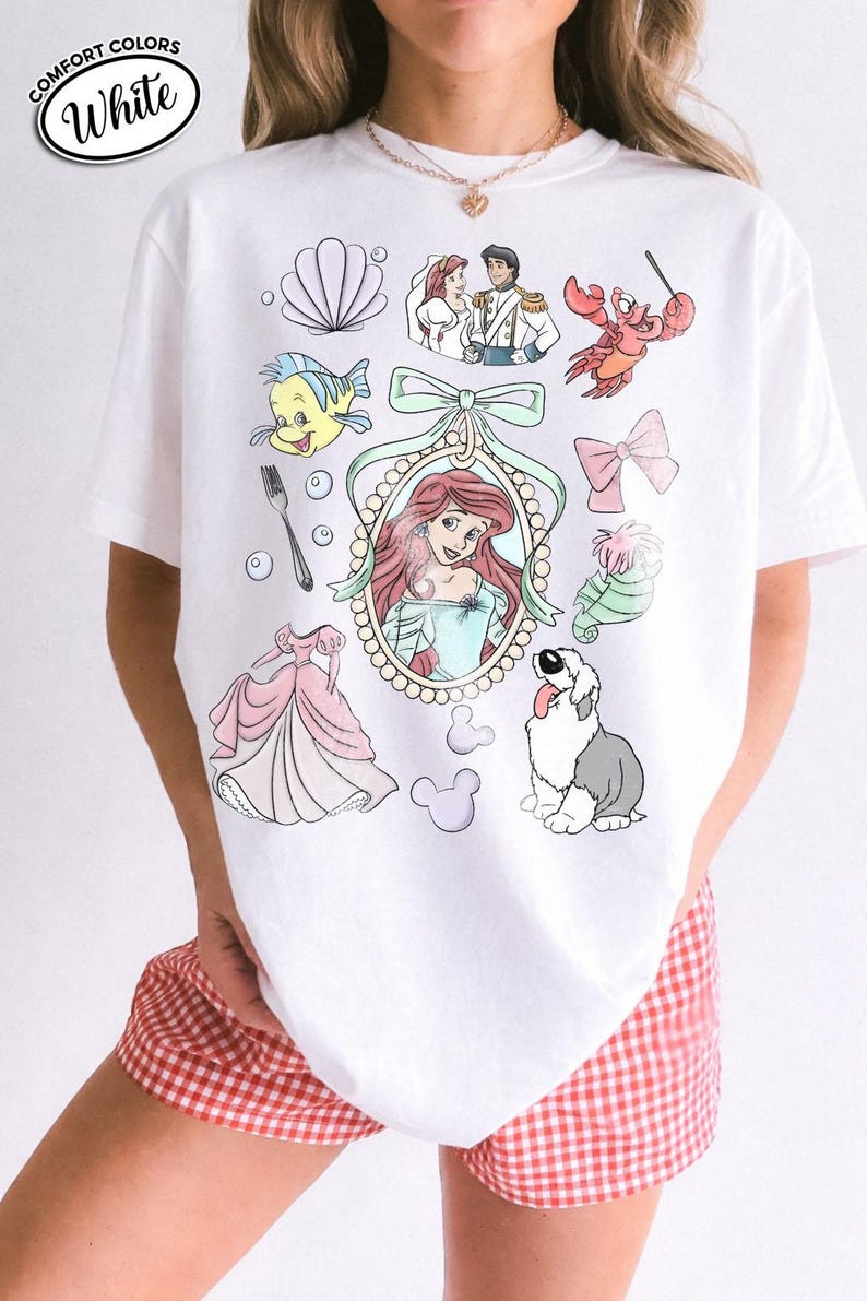 May include: White t-shirt with a colorful graphic featuring characters and elements from a popular animated movie. The design includes a princess, a crab, a fish, and other related imagery. The shirt has the words "Comfort Colors" and "White" in a circle.