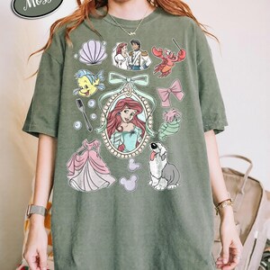 May include: Moss green t-shirt featuring a collection of cartoon illustrations. The design includes a portrait of a red-haired woman, a pink dress, a dog, and other characters. The words "Comfort Colors" and "Moss" are in a white oval.