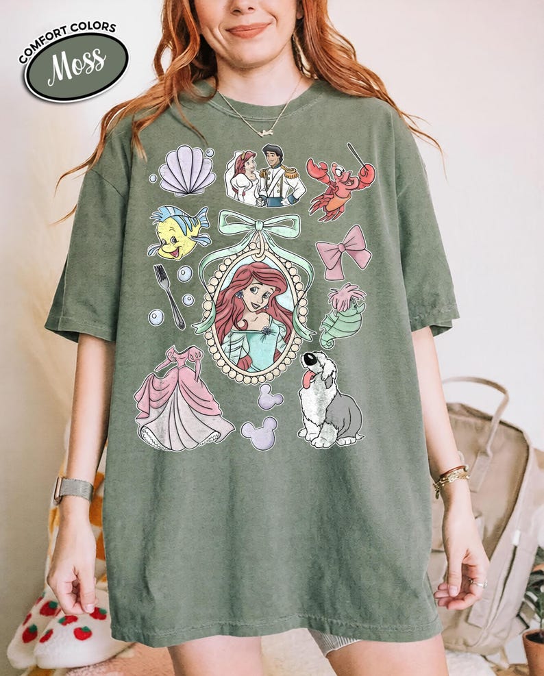 May include: Moss green t-shirt featuring a collection of cartoon illustrations. The design includes a portrait of a red-haired woman, a pink dress, a dog, and other characters. The words "Comfort Colors" and "Moss" are in a white oval.