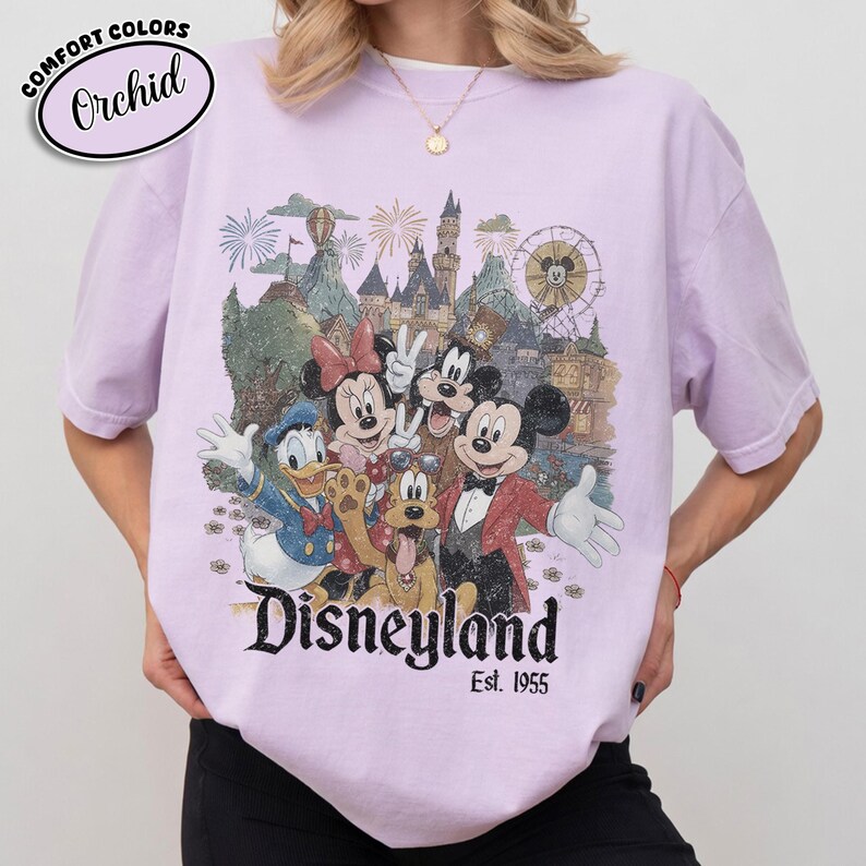 Może przedstawiać: Lawendowa koszulka Orchid Comfort Colors z grafiką Disneyland. Projekt przedstawia Myszka Miki, Minnie, Kaczora Donalda, Goofy'ego i Pluto, a także zamek, fajerwerki i diabelski młyn. Tekst "Disneyland Est. 1955" jest wydrukowany poniżej.