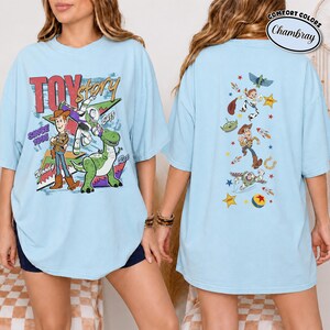 K&ouml;nnte beinhalten: Ein hellblaues T-Shirt mit Toy Story-Grafiken. Die Vorderseite zeigt Woody, Buzz Lightyear und Rex mit dem Text "TOY story SINCE 1995". Die R&uuml;ckseite zeigt eine vertikale Anordnung von Charakteren, darunter Woody, Buzz und ein Alien mit Sternen.
